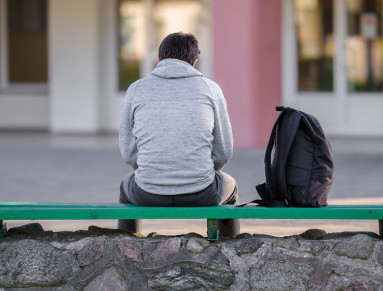 Un adolescent vu de dos est assis sur un banc devant un lycée.