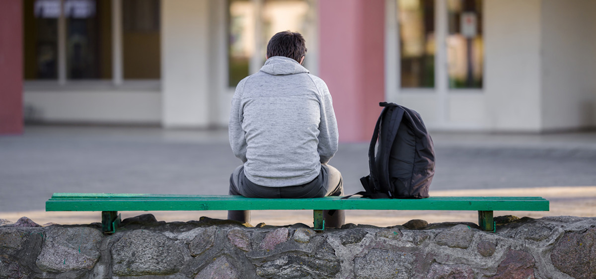 Un adolescent vu de dos est assis sur un banc devant un lycée.